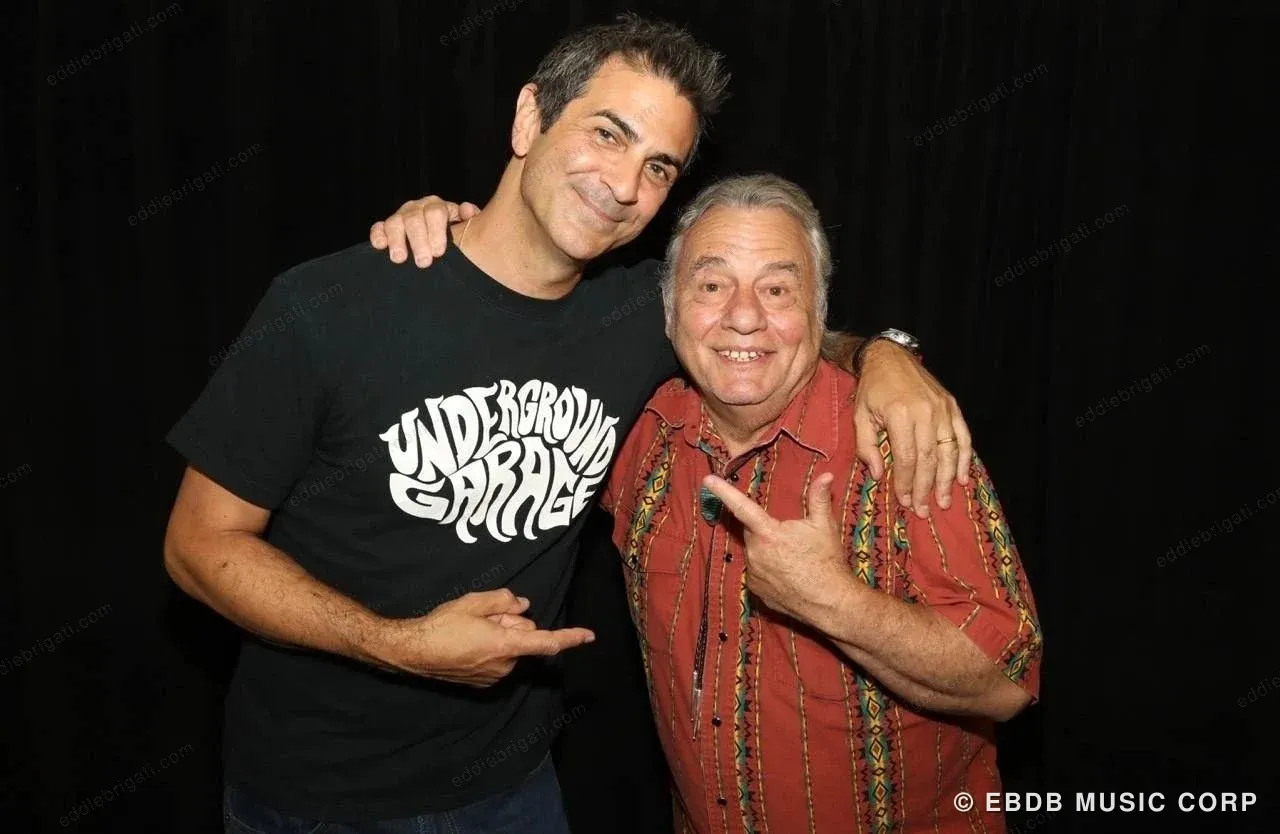 Two men with their arms around each other, laughing — one in a black graphic T-shirt, the other in a red patterned shirt.