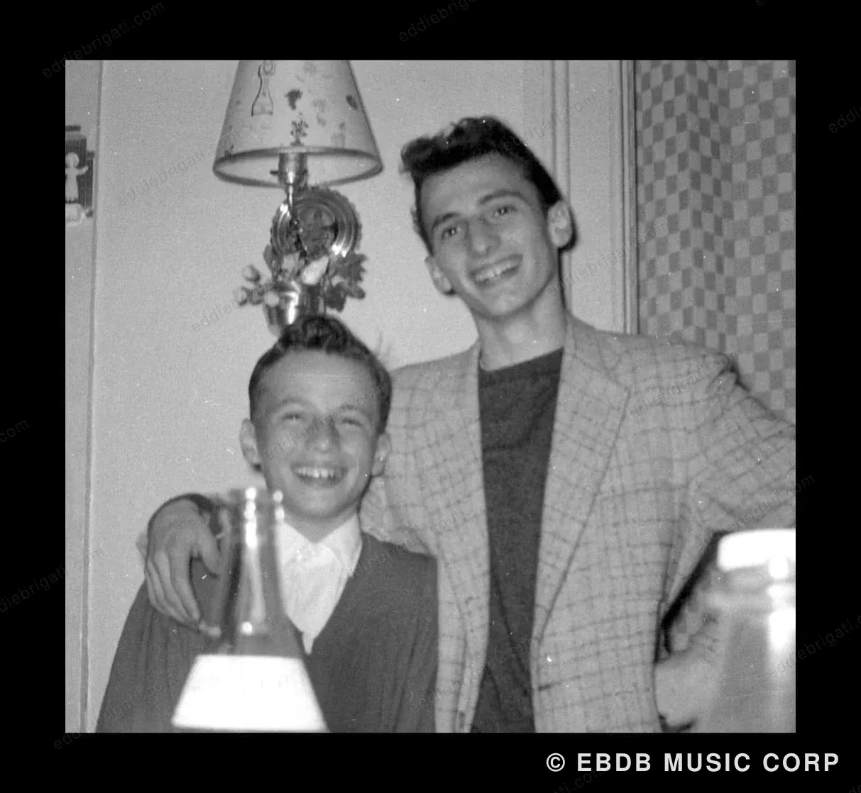 A black-and-white snapshot of two Brigati brothers as boys at home, one holding a small trophy or lamp base, both smiling in front of patterned wallpaper.