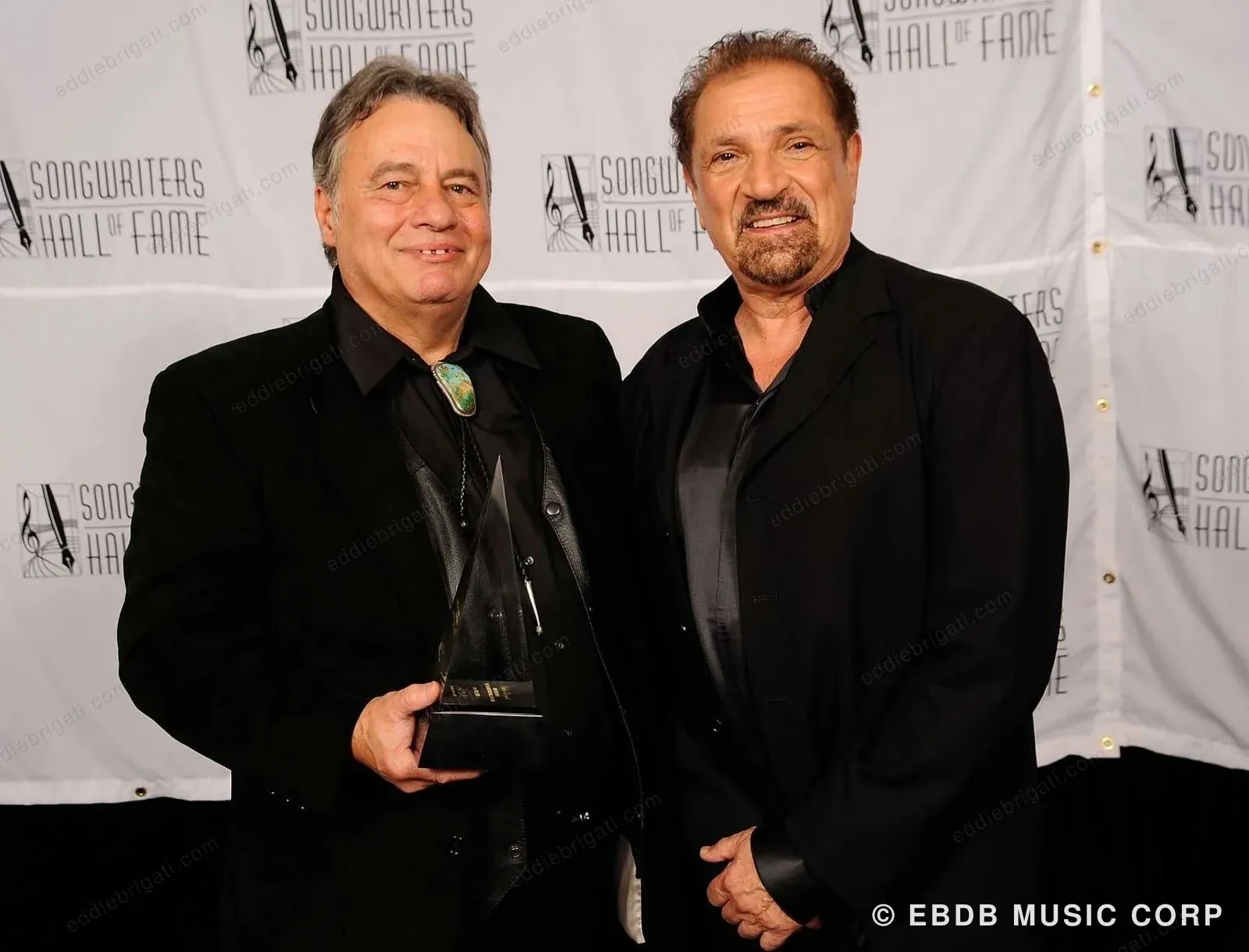 Eddie Brigati and Felix Cavaliere standing together in black, in front of a Songwriters Hall of Fame step-and-repeat, Eddie holding an award.