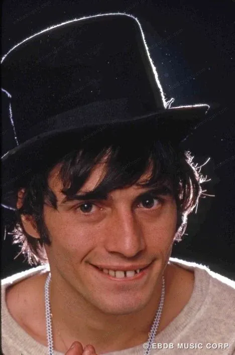 A colour photograph of a young Eddie Brigati in a black top hat, smiling at the camera.