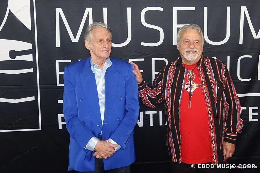 Two men standing together in front of a Grammy Museum Experience sign, one in a blue blazer, the other in a red and black patterned shirt with a bolo tie.