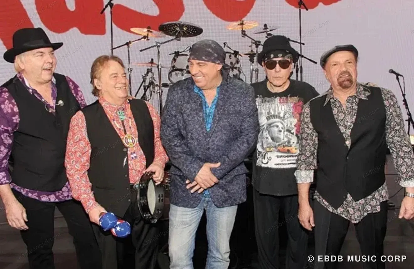 Five men standing together on stage in front of a drum kit and a large red “Rascals” backdrop, in waistcoats and hats.