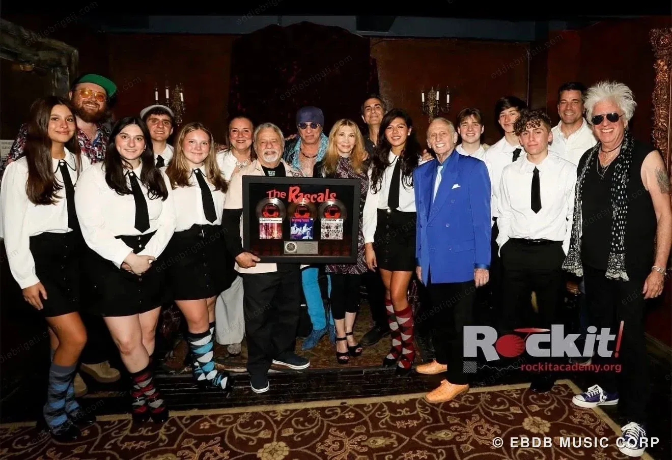 A large group of Rockit Academy students and adults posed together indoors, two of them holding a framed Rascals presentation award.