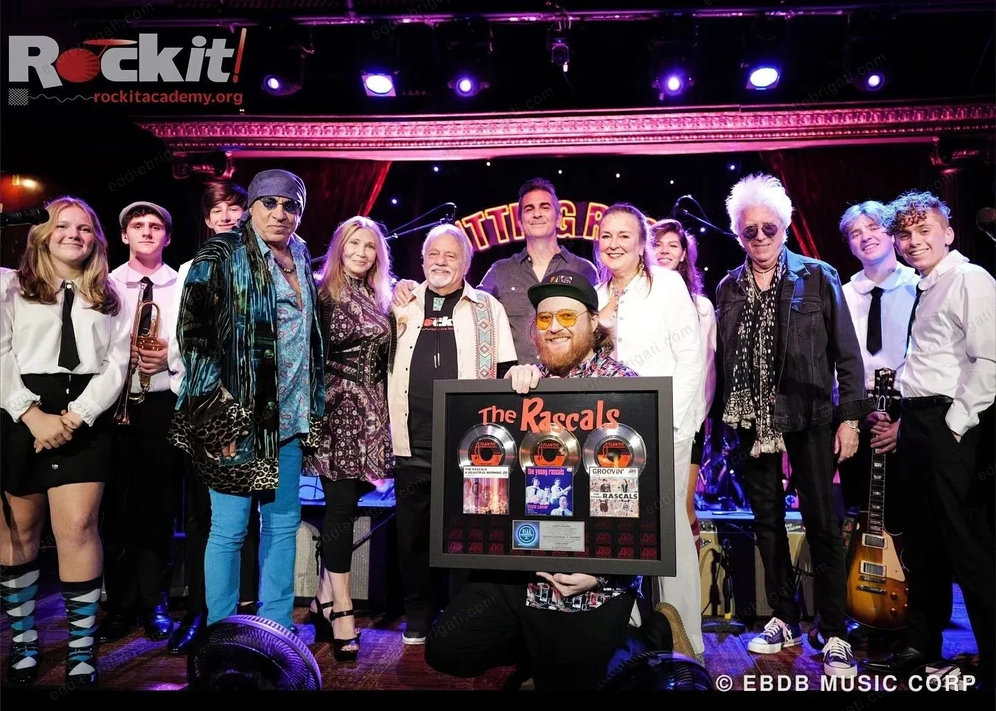 Rockit Academy students and guests on a stage under a Rockit banner, holding a framed Rascals presentation award.